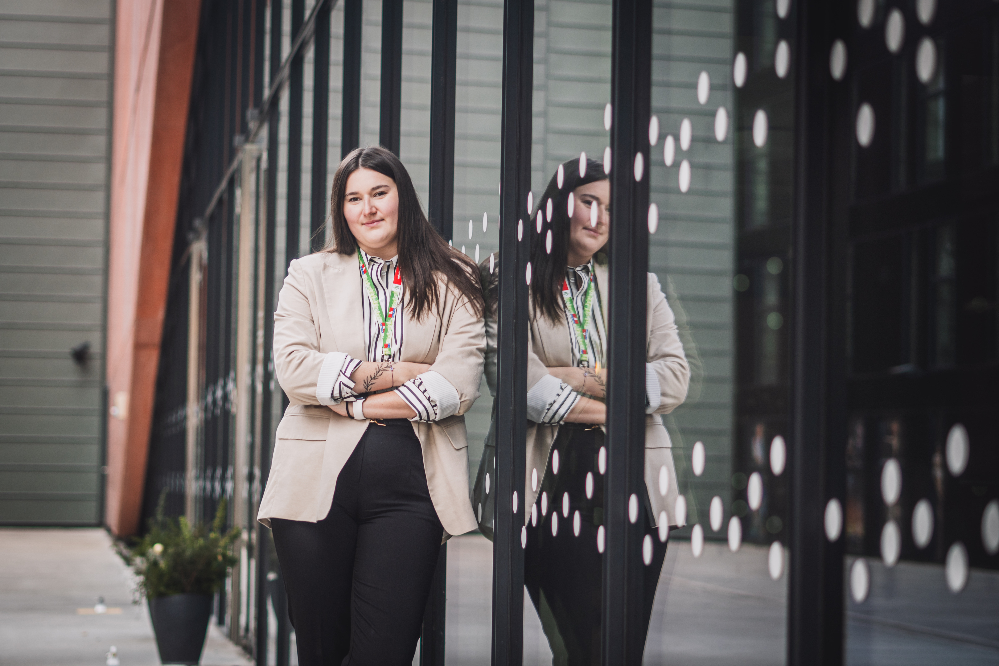 Stylový business portrét mladé ženy v saku, ležérně opřené o moderní fasádu s jejím odrazem ve skle.