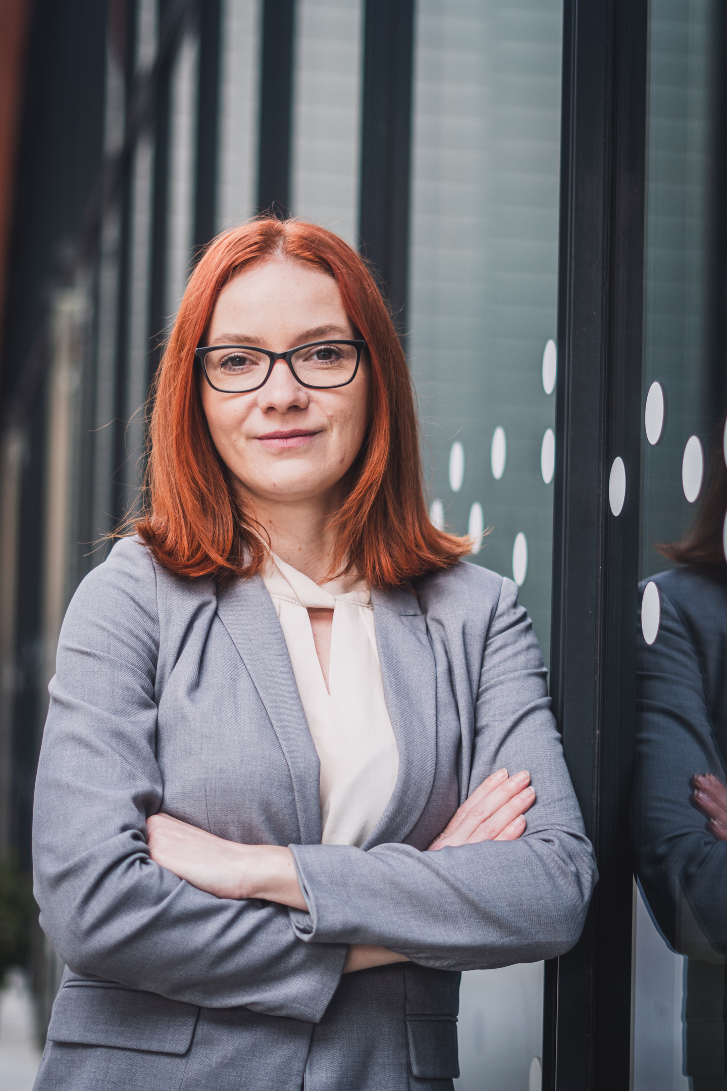 Profesionální headshot ženy s brýlemi a zrzavými vlasy v kostýmku, s jemným úsměvem a odrazem ve skle.