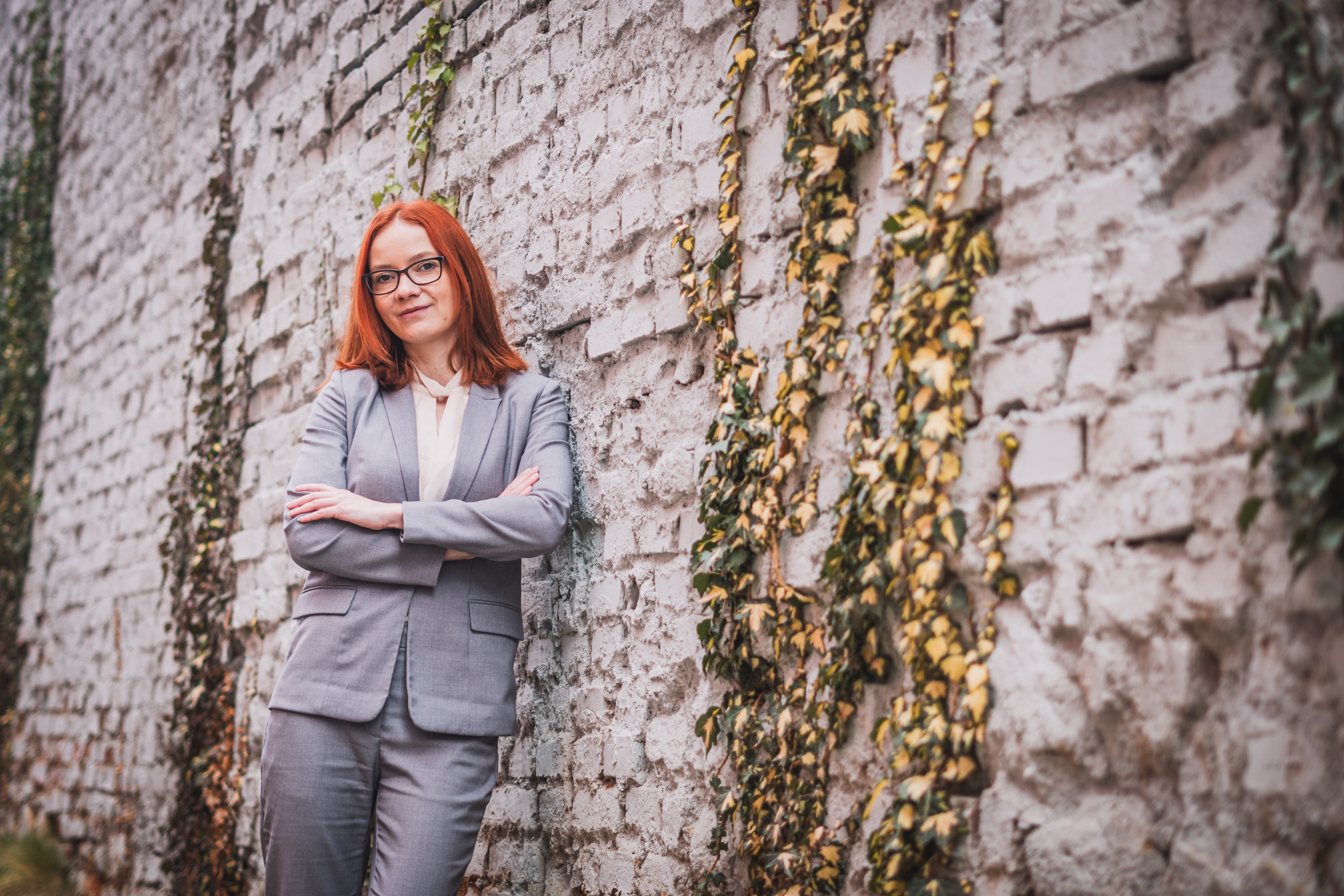 Elegantní manažerský portrét ženy v šedém kostýmku, opírající se o starou cihlovou zeď s popínavými rostlinami.