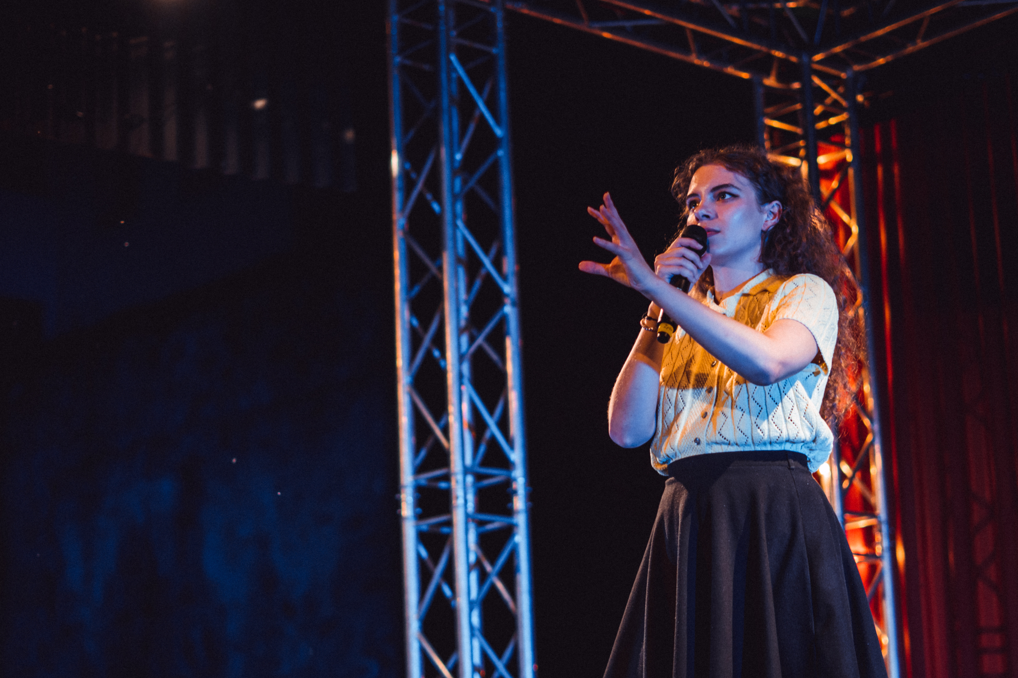 Účinkující žena s kudrnatými vlasy gestikuluje a mluví do mikrofonu během svého vystoupení na slam poetry večeru.