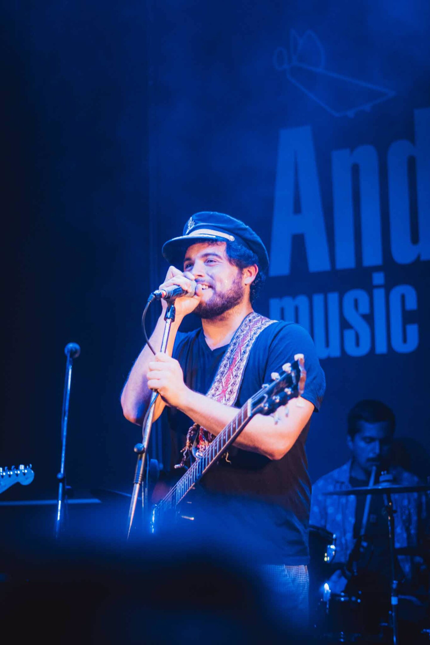 Zpěvák s kytarou a kapitánskou čepicí energicky zpívá na pódiu v modrém světle během hudebního vystoupení.