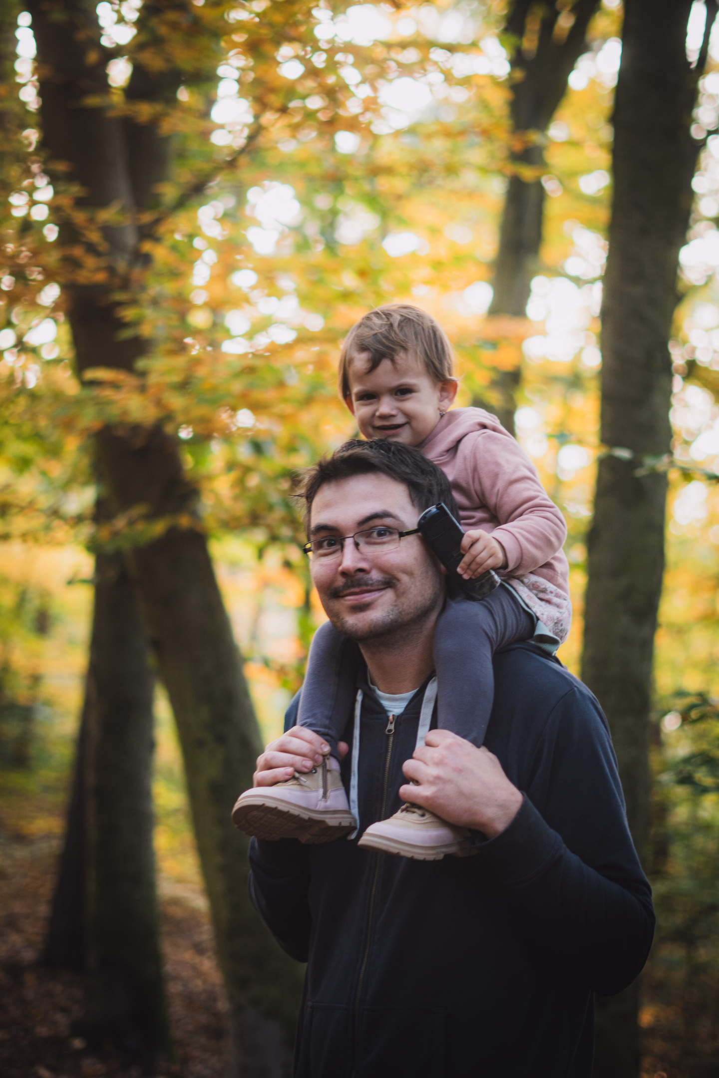 Přirozený portrét otce a dítěte na ramenou během rodinného focení v parku se zlatavým podzimním světlem.