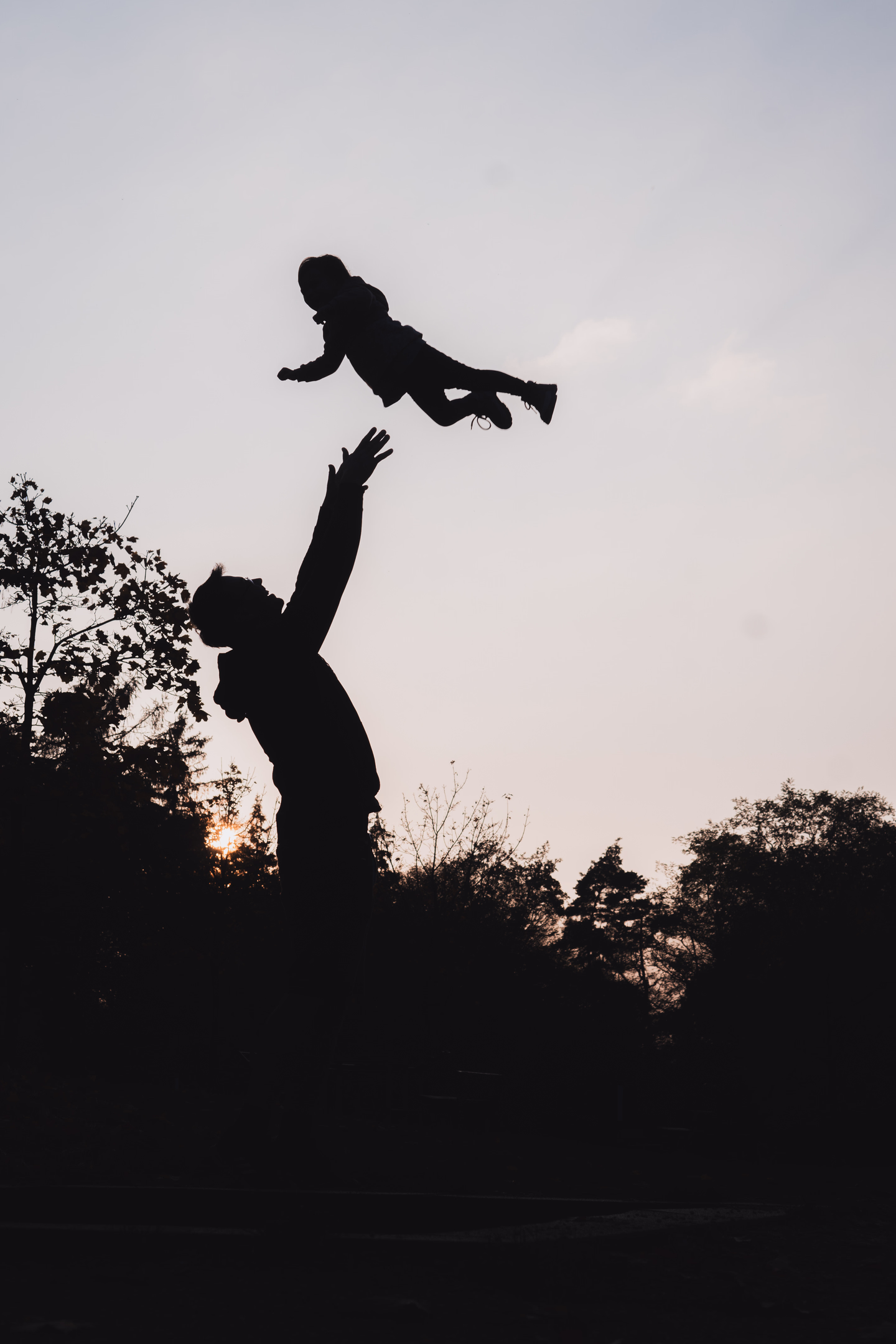 Silueta otce vyhazujícího dítě do vzduchu při západu slunce, zachycující radostný a hravý moment.