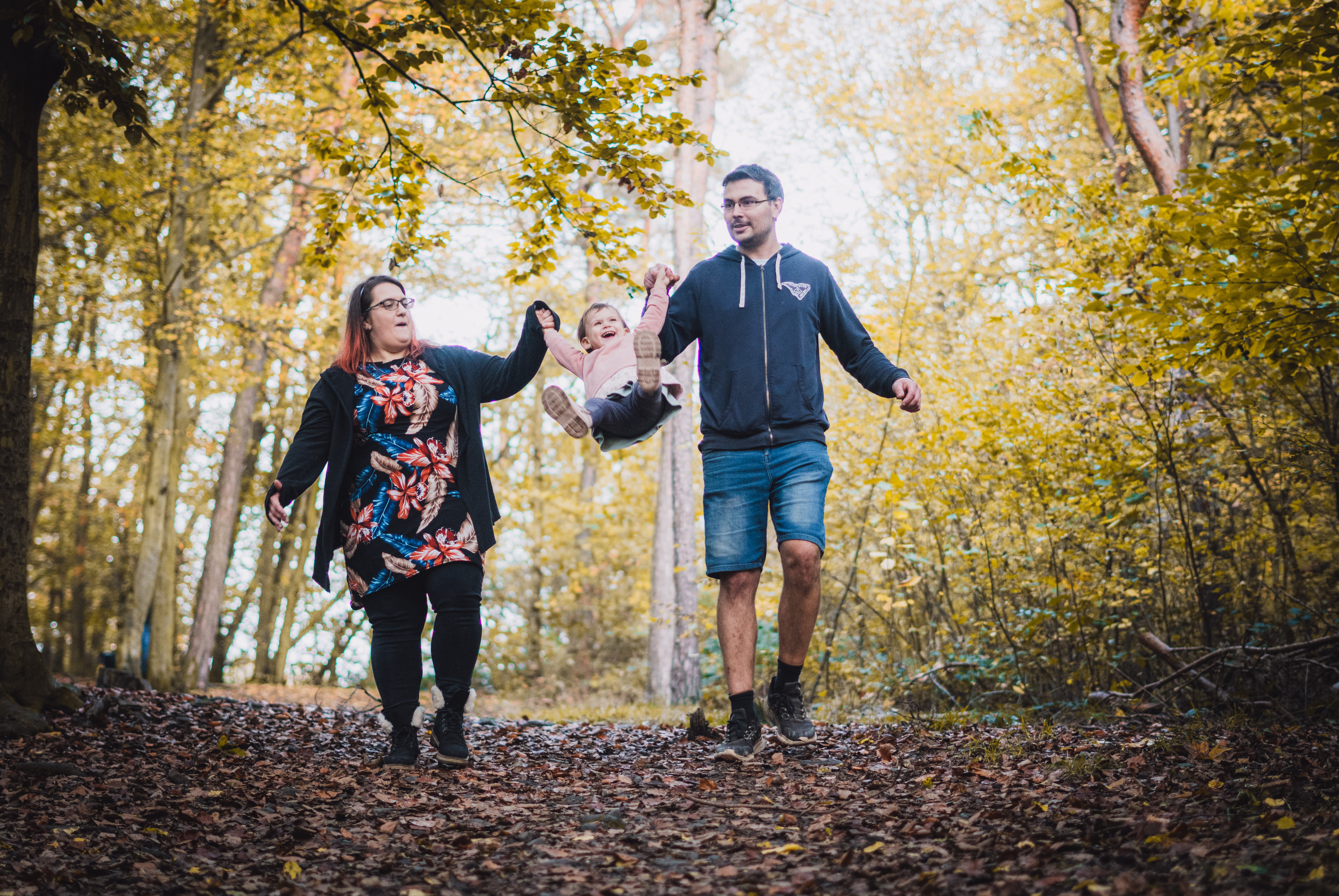 Rodina s malou holčičkou se prochází podzimním lesem a rodiče ji vesele houpu mezi sebou.