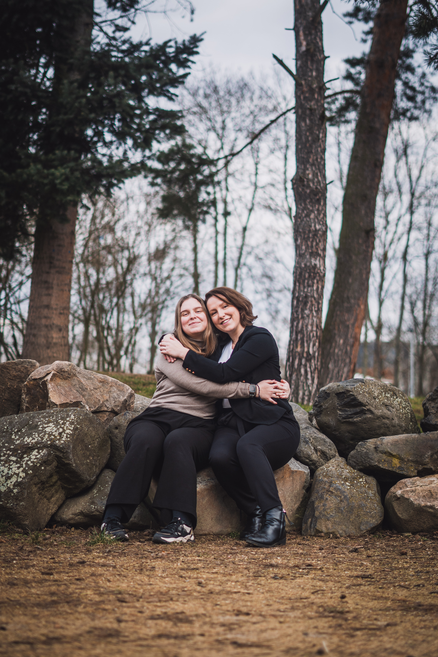 Portrét dvou žen, matky a dcery, které se usmívají a objímají sedící na kamenech v parku.