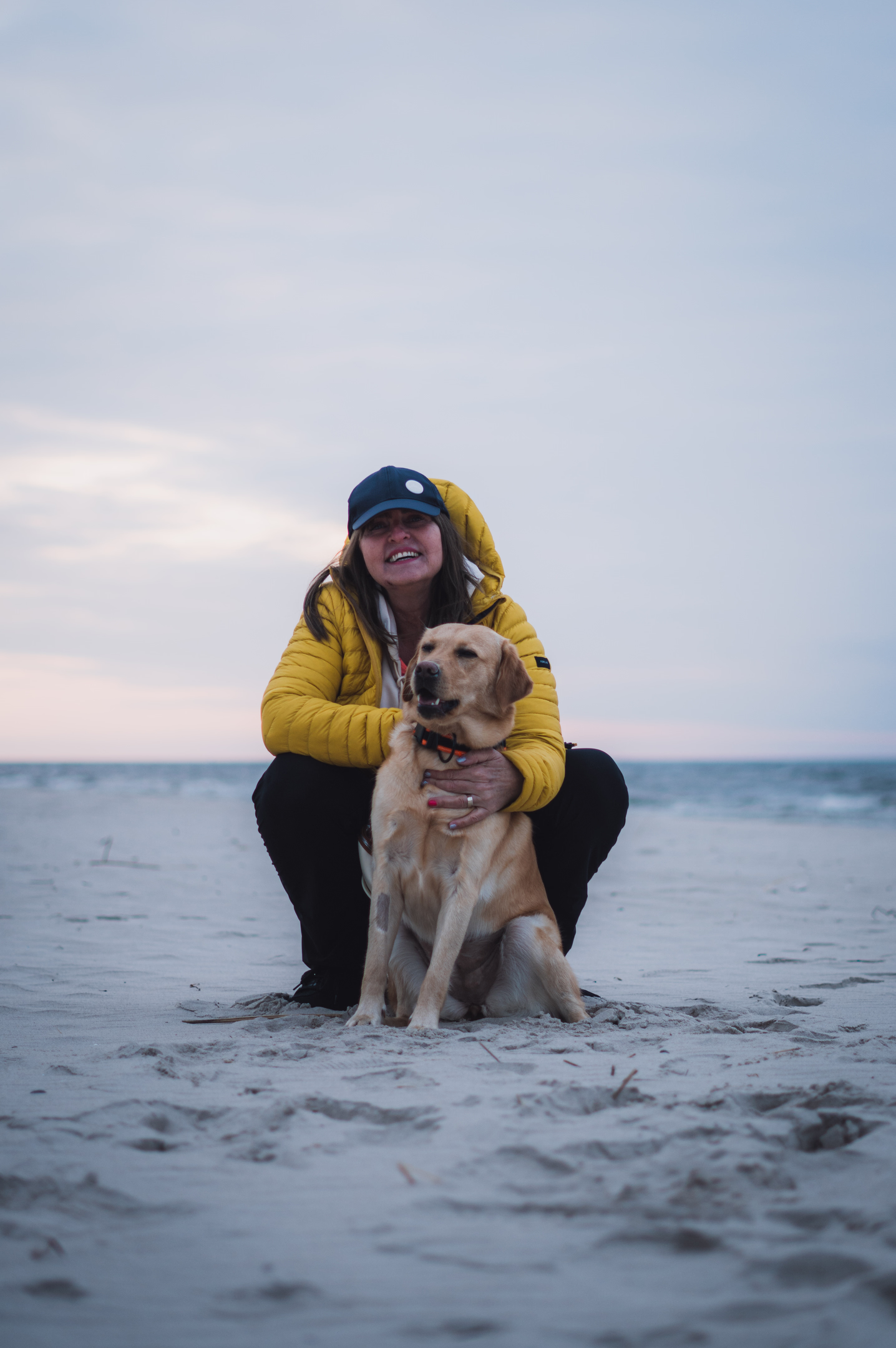 Usměvavá žena v podřepu objímá svého labradora na písečné pláži během focení mazlíčků.